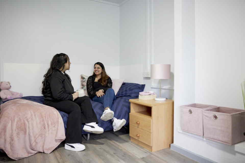 2 female students sat talking on a bed.